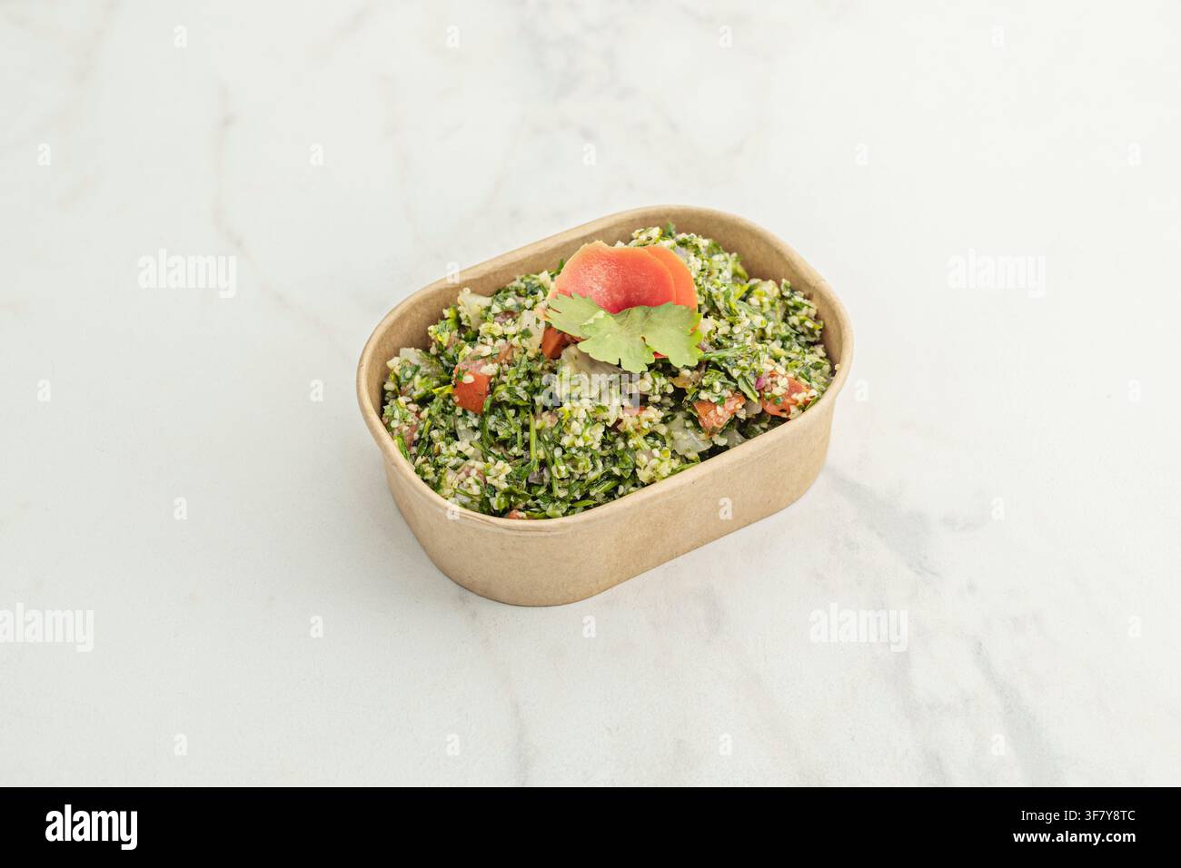 Taboulé mit Bulgur, Tomate und Koriander-Garnitur in einem Take-out-Behälter aus Kraftpapier auf Marmor. Stockfoto