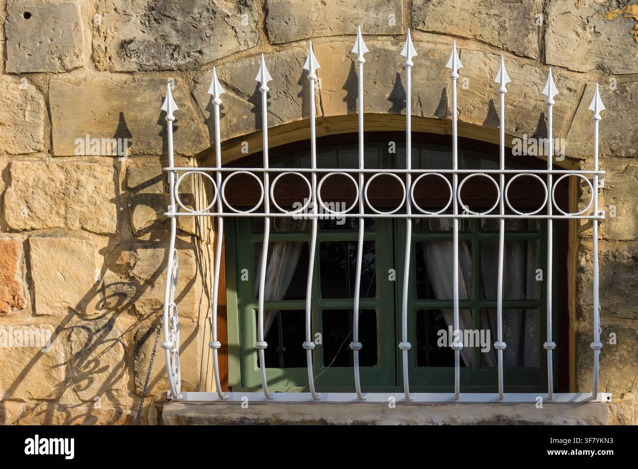 Grünes gewölbtes Fenster mit dekorativen weißen schmiedeeisernen Gittern an einer rustikalen Steinwand im Sonnenlicht Stockfoto