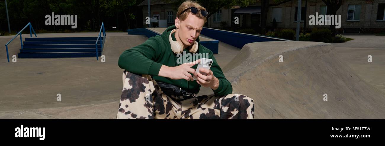 Attraktiver junger Mann sitzt bequem im Skatepark und konzentriert sich auf sein Handy inmitten der lebendigen Szene. Stockfoto