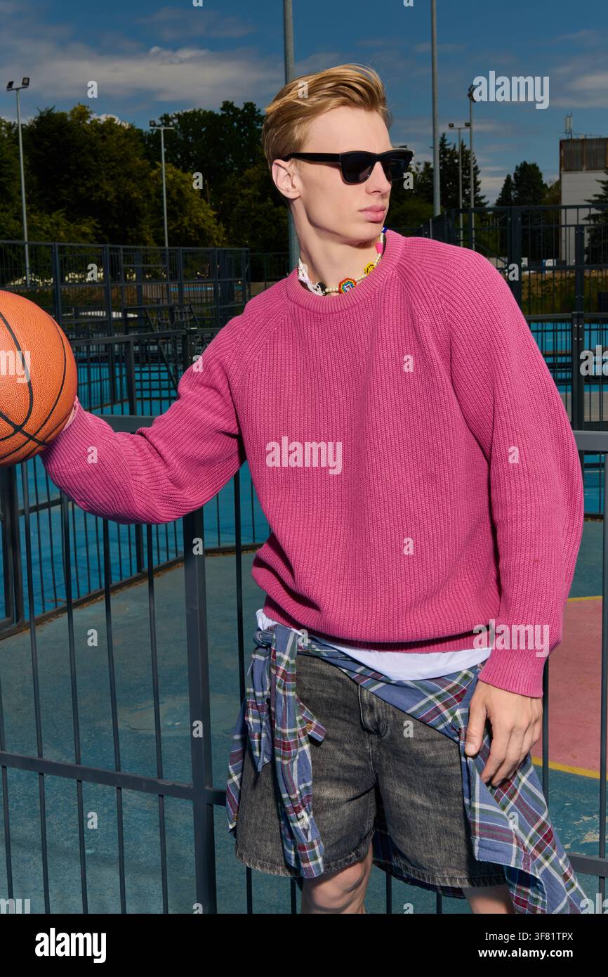 Junger Mann in einem rosa Pullover posiert mit einem Basketball und strahlt Coolness im Park aus. Stockfoto