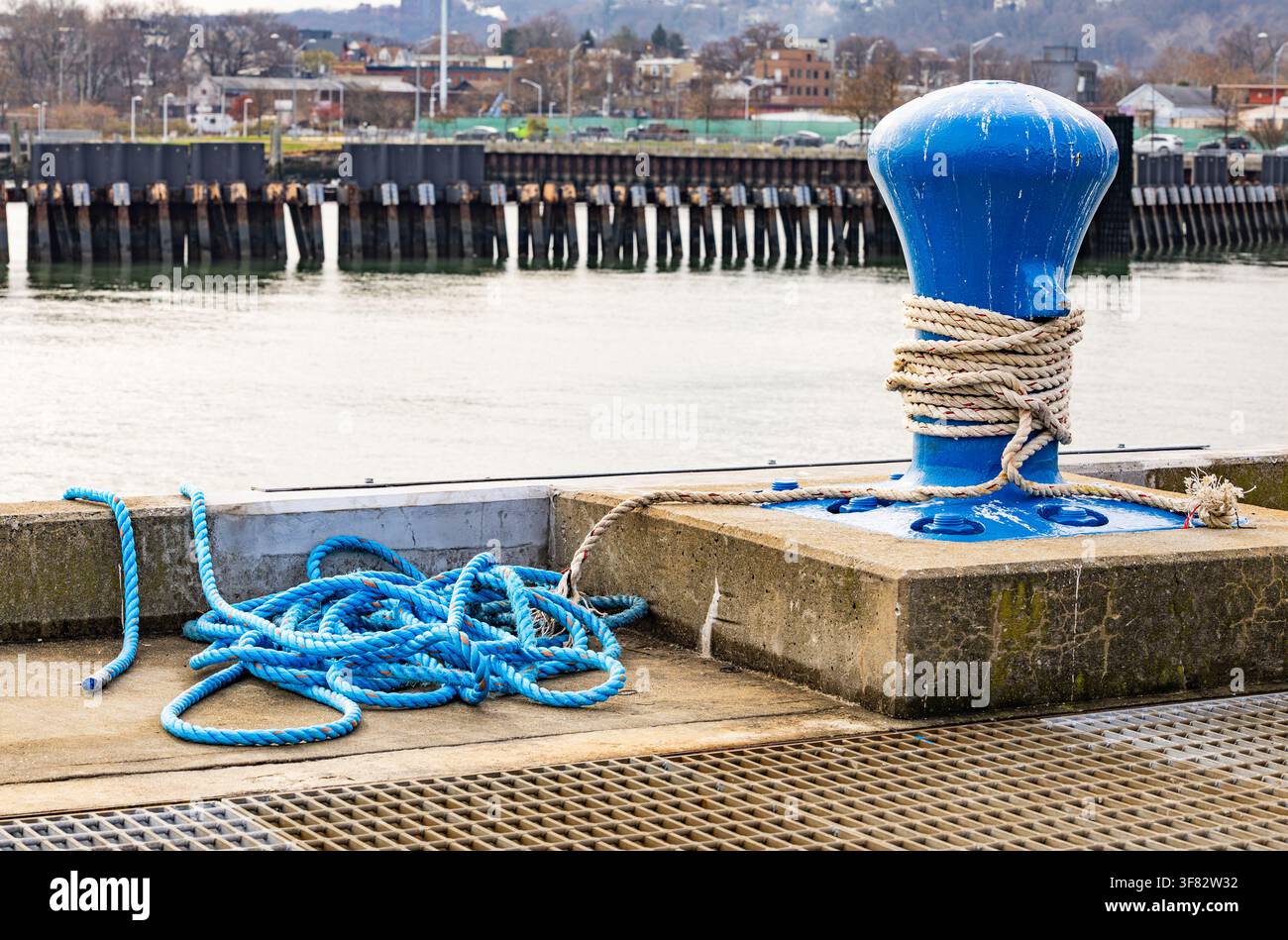 Hellblauer Ankerpfahl aus Gusseisen, mit Seil umwickelt und verheddertem blauem Schnur an einem Betonpier im Marinehafen mit Wasserhintergrund Stockfoto