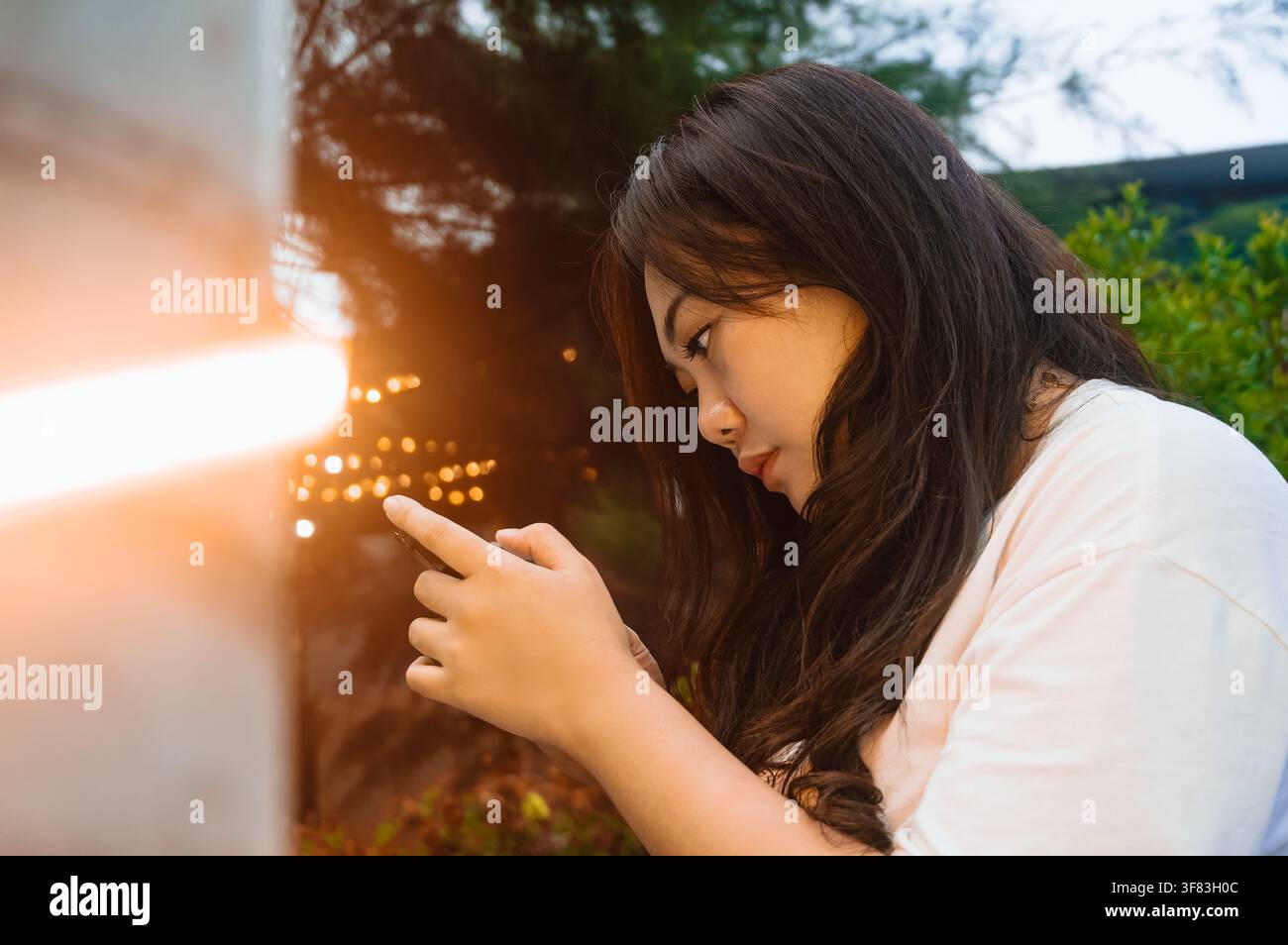 Junge Frau, die abends im Freien ein Smartphone benutzt, von der Seite betrachtet, mit dekorativen Lichtern, die eine warme und stimmungsvolle Lebensstil-Szene erzeugen. Stockfoto