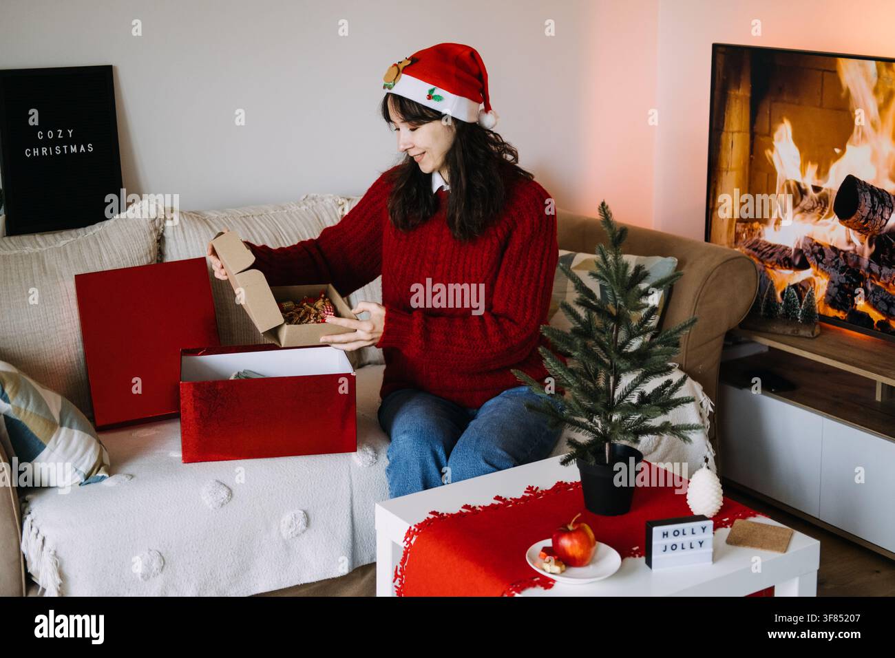 Frau mit Weihnachtsmütze öffnet Kiste mit Ornamenten auf dem Sofa neben den Schildern COZY CHRISTMAS und HOLLY JOLLY. Auspacken von Weihnachtsdekorationen, Auspacken von Feiertagsdekorationen Stockfoto