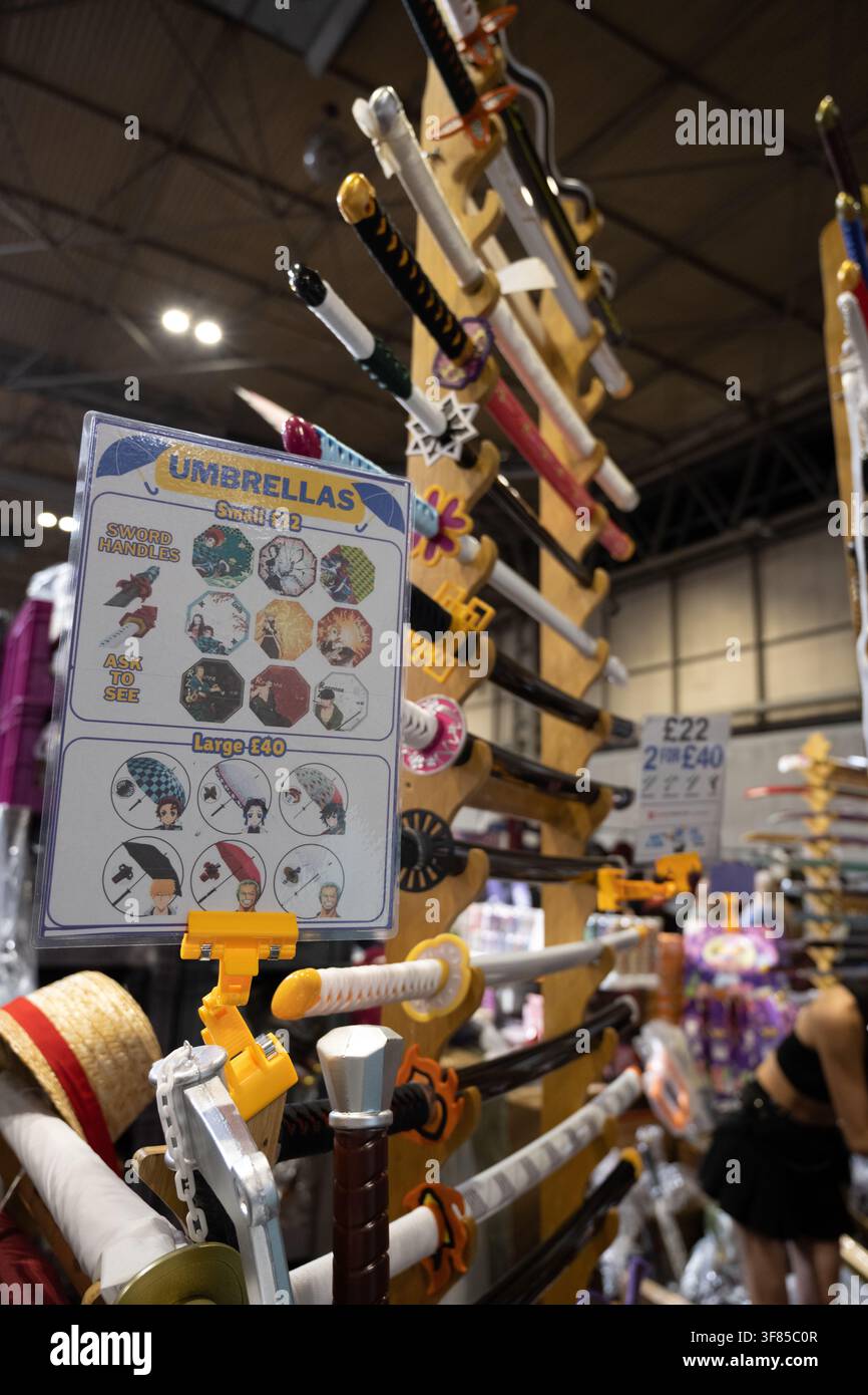 Eine Ausstellung von Anime-inspirierten Merchandisingartikeln, darunter dekorative Ausstellungsstäbe auf vertikalen Holzständern und ein Schild, das thematische Regenschirme mit schwertähnlichen Griffen bewirbt, an einem Händlerstand während der Birmingham MCM Comic Con im NEC am 8. August 2026. Stockfoto