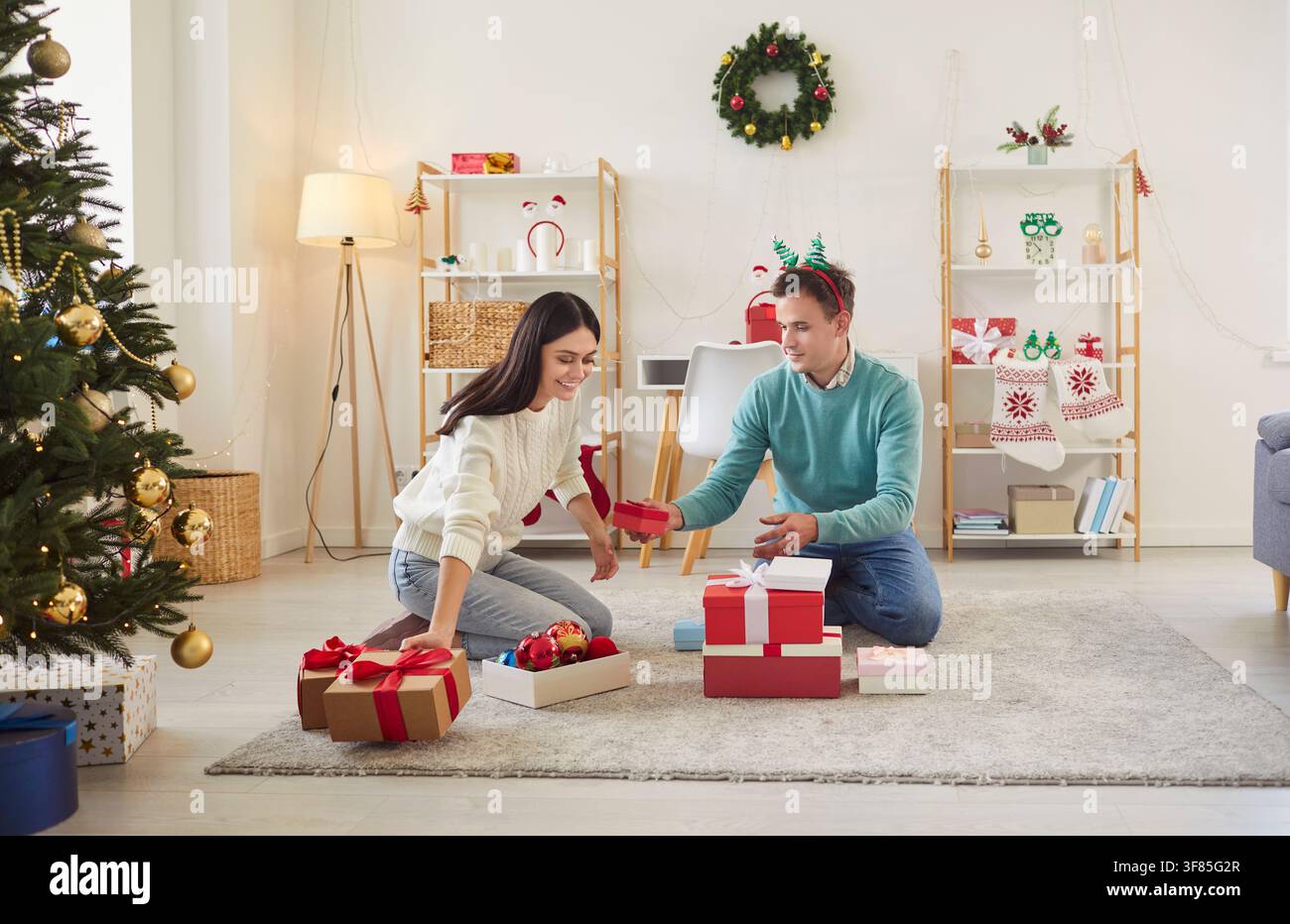 Fröhliches Paar sitzt auf einem Teppich in ihrem gemütlichen Wohnzimmer und tauscht an Weihnachten Geschenke aus. Stockfoto