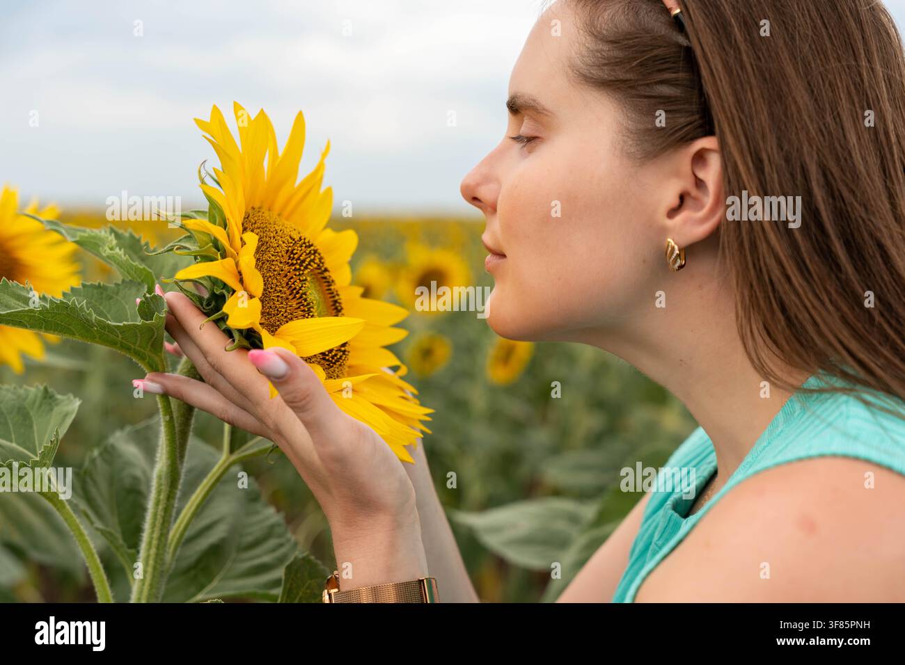 Junge Frau mit geschlossenen Augen lehnt sich zu einer hellen Sonnenblume in einem blühenden Feld, aufgenommen im Seitenprofil als Nahaufnahme mit weichem natürlichem Licht Stockfoto