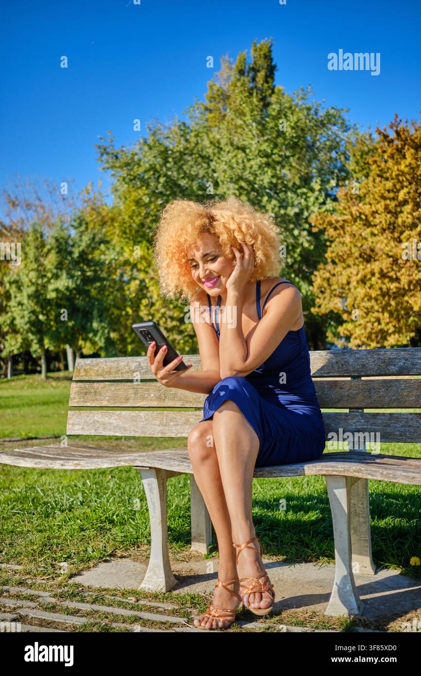 Frau lächelt und interagiert mit ihrem Mobiltelefon, während sie auf einer Parkbank sitzt Stockfoto