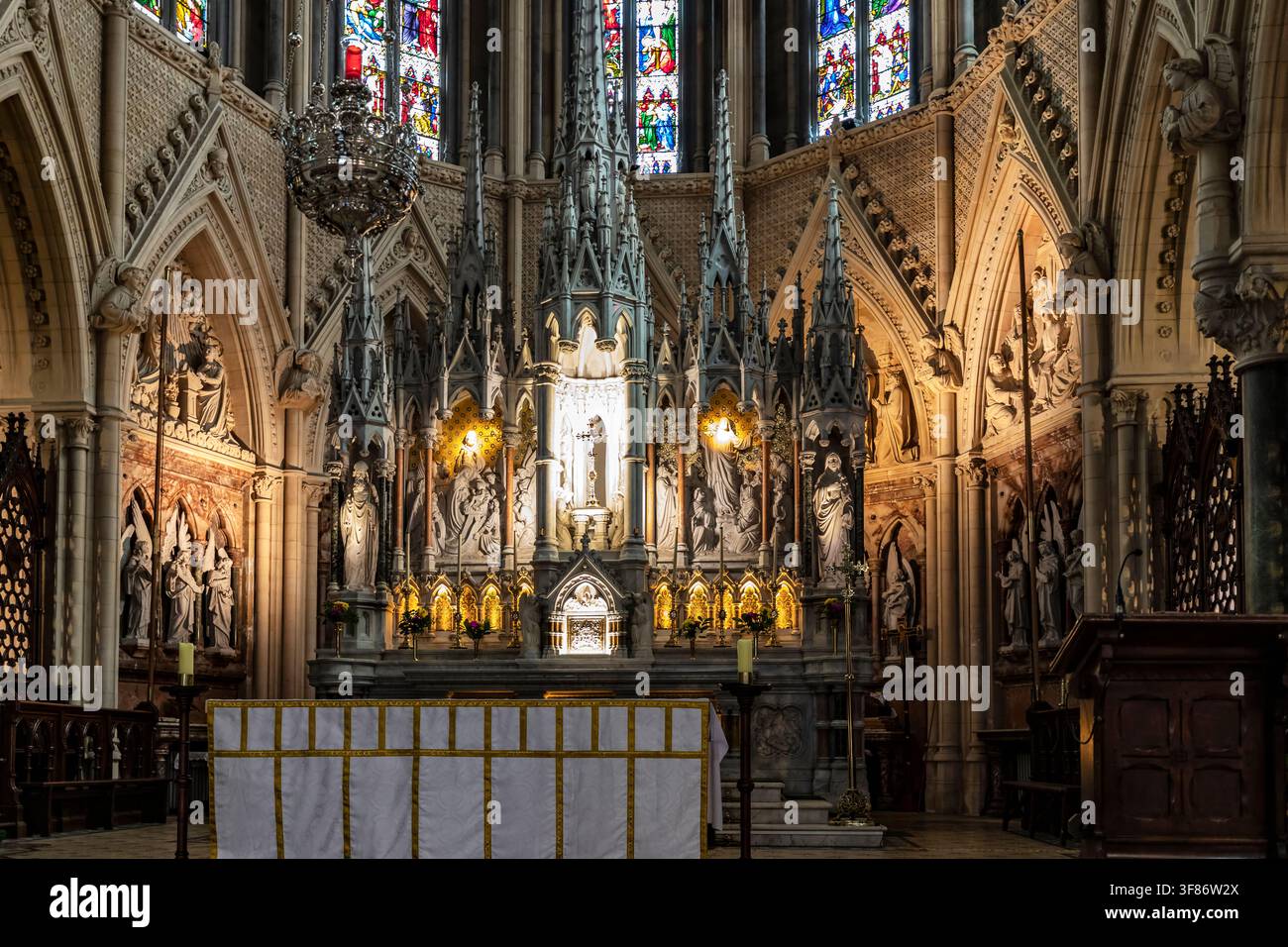 Verzierter neugotischer Hochaltar und geschnitzter Rückwandaltar unter bunten Buntglasfenstern in St. Colmans Kathedrale, Cobh, County Cork, Irland Stockfoto