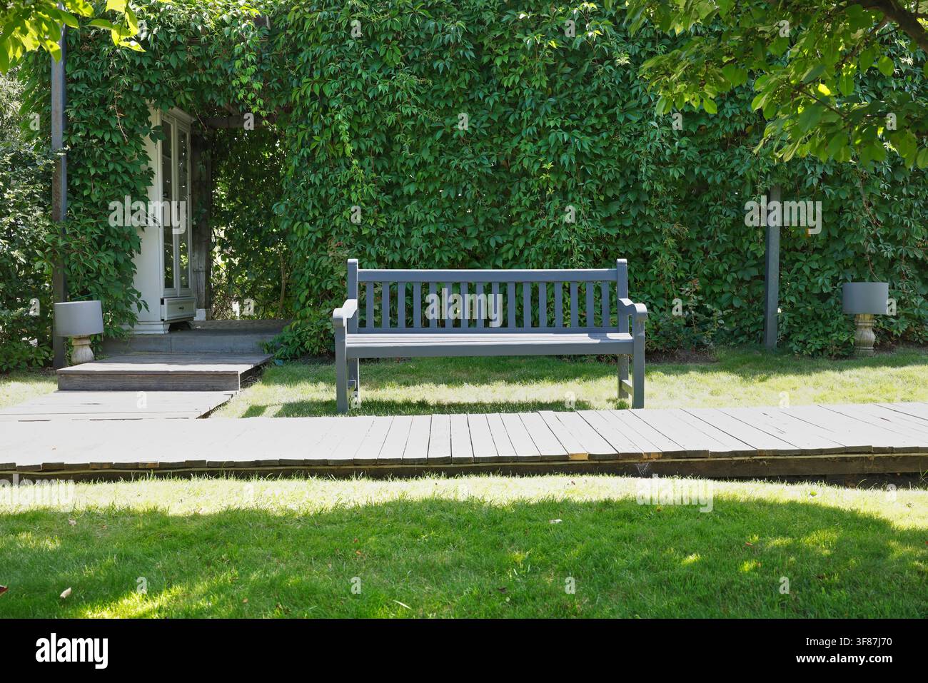 Holzbank in einer ruhigen grünen Gartenecke. Gemütliches, nachhaltiges Sitzplatzdesign für die Hinterhofterrasse. Friedlicher Sommerparkweg als Naturhintergrund. Stockfoto
