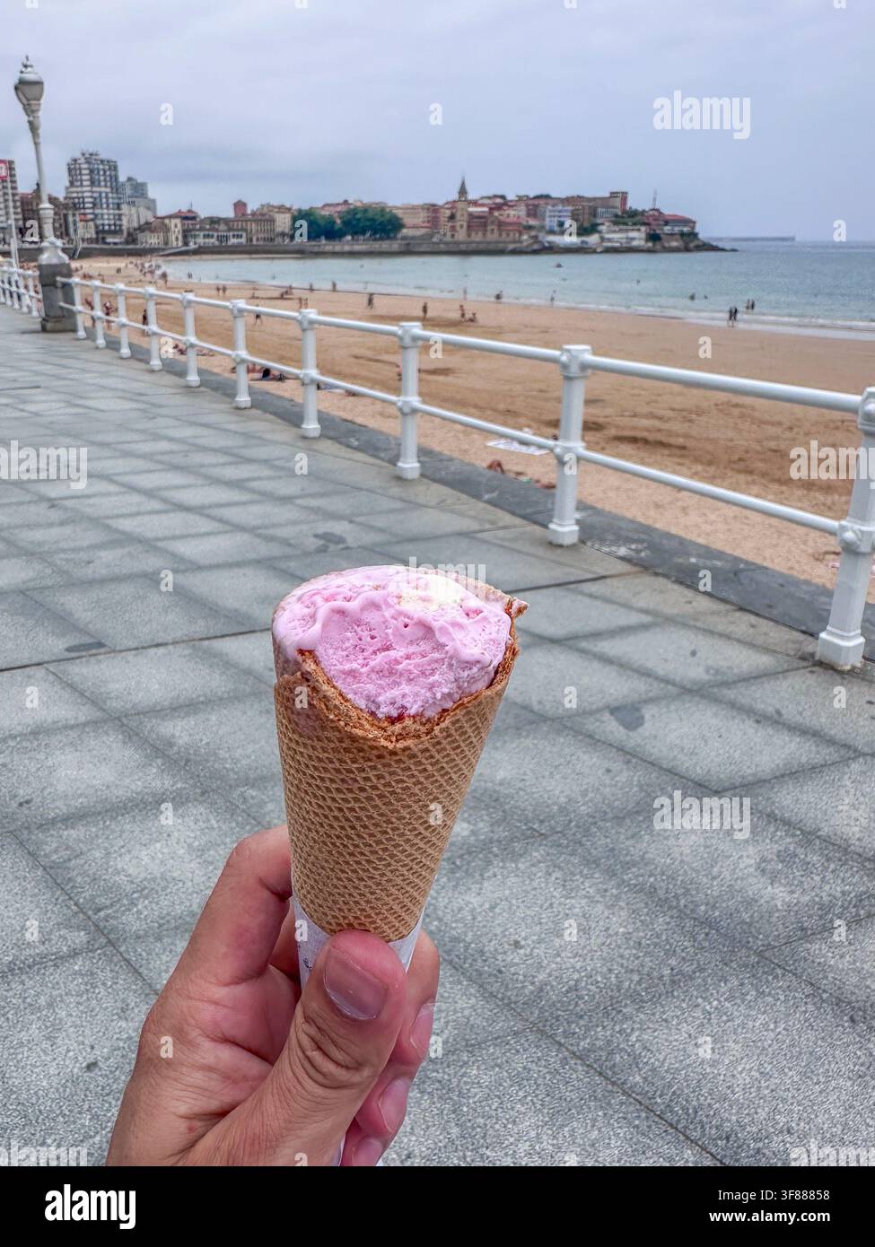 Eine Hand hält eine Eiswaffel mit einer rosa Kugel auf der Strandpromenade von San Lorenzo in Gijón, Asturien, Spanien Stockfoto