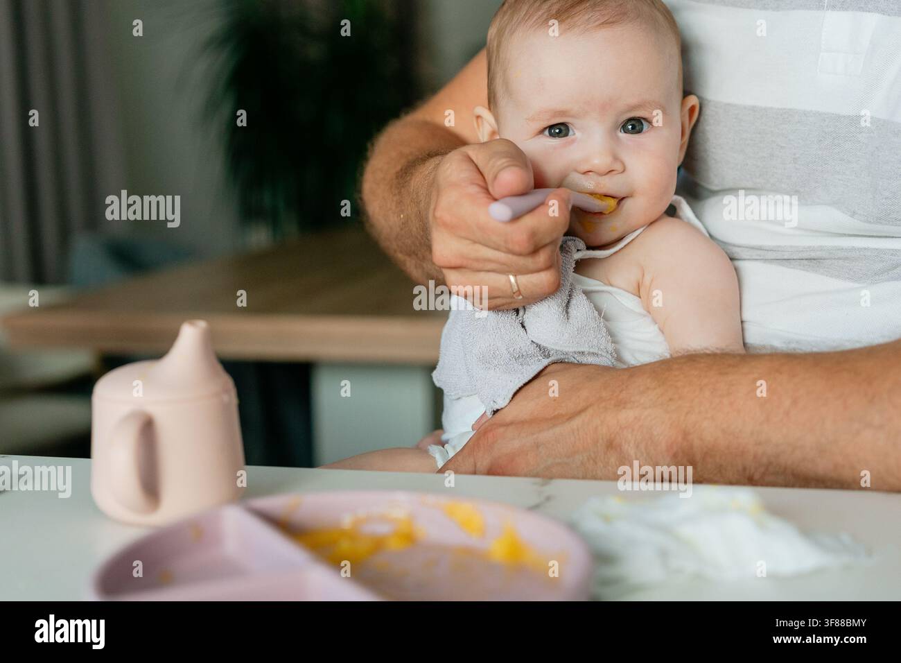 eine zarte Szene eines Babys, das mit einem Löffel gefüttert wird, in einer warmen häuslichen Umgebung, die die fürsorglichen und liebevollen Aspekte des Familienlebens hervorhebt Stockfoto