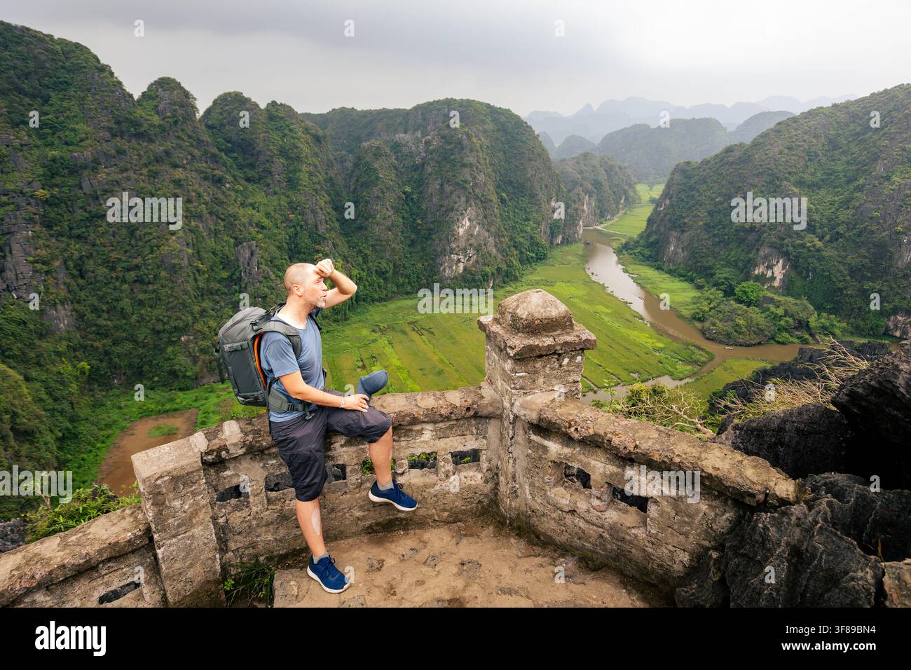 Tourist mit Rucksack macht eine Pause auf einer Felsklippe während einer heißen Tageswanderung in den Kalkstein-Karstbergen in Ninh Binh, Vietnam. Reisfelder, windig Stockfoto