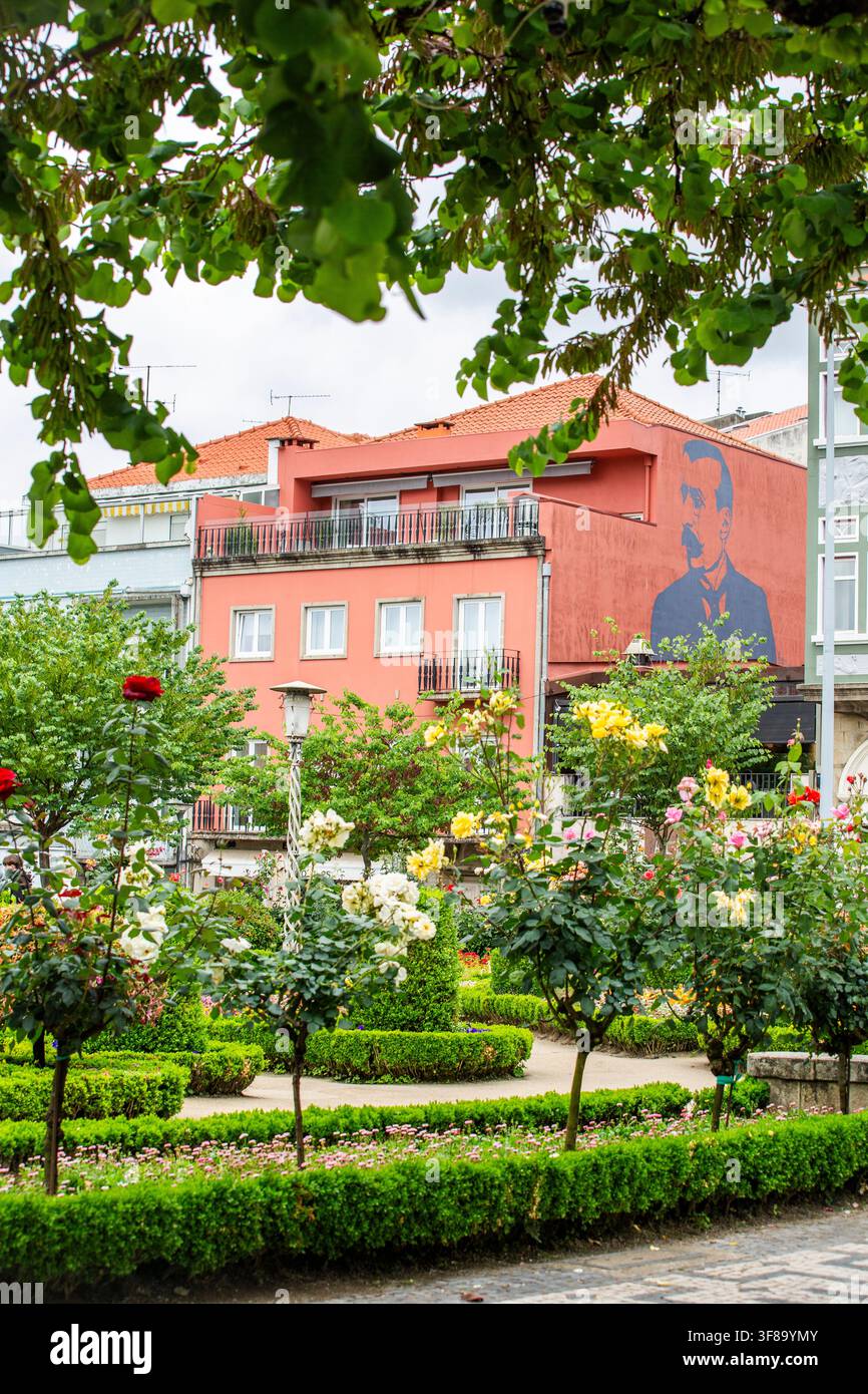 Lebendiger Garten mit üppiger Vegetation und blühenden Blumen. Ein Gebäude mit rotem Dach steht hinter gepflegten Hecken und Bäumen. Ein Wandgemälde eines Mannes schmückt die Seite Stockfoto