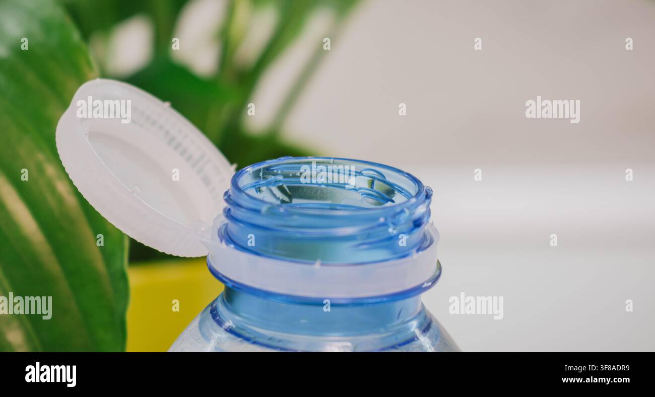 Makroaufnahme einer offenen aufgesetzten Kappe auf einer blauen Plastik-Wasserflasche in der Nähe von grünen Pflanzenblättern, umweltfreundliches Design, EU-Richtlinie zum befestigten Verschluss, Stockfoto Stockfoto