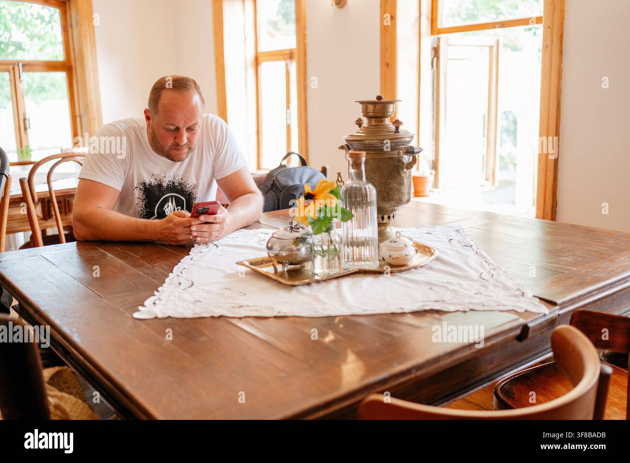 Mann, der auf ein Telefon schaut, sitzt drinnen in der Nähe eines Vintage-Samowars auf einer Spitzendecke. Perfektes Stockbild für den Kontrast zwischen traditioneller Teekultur und Stockfoto