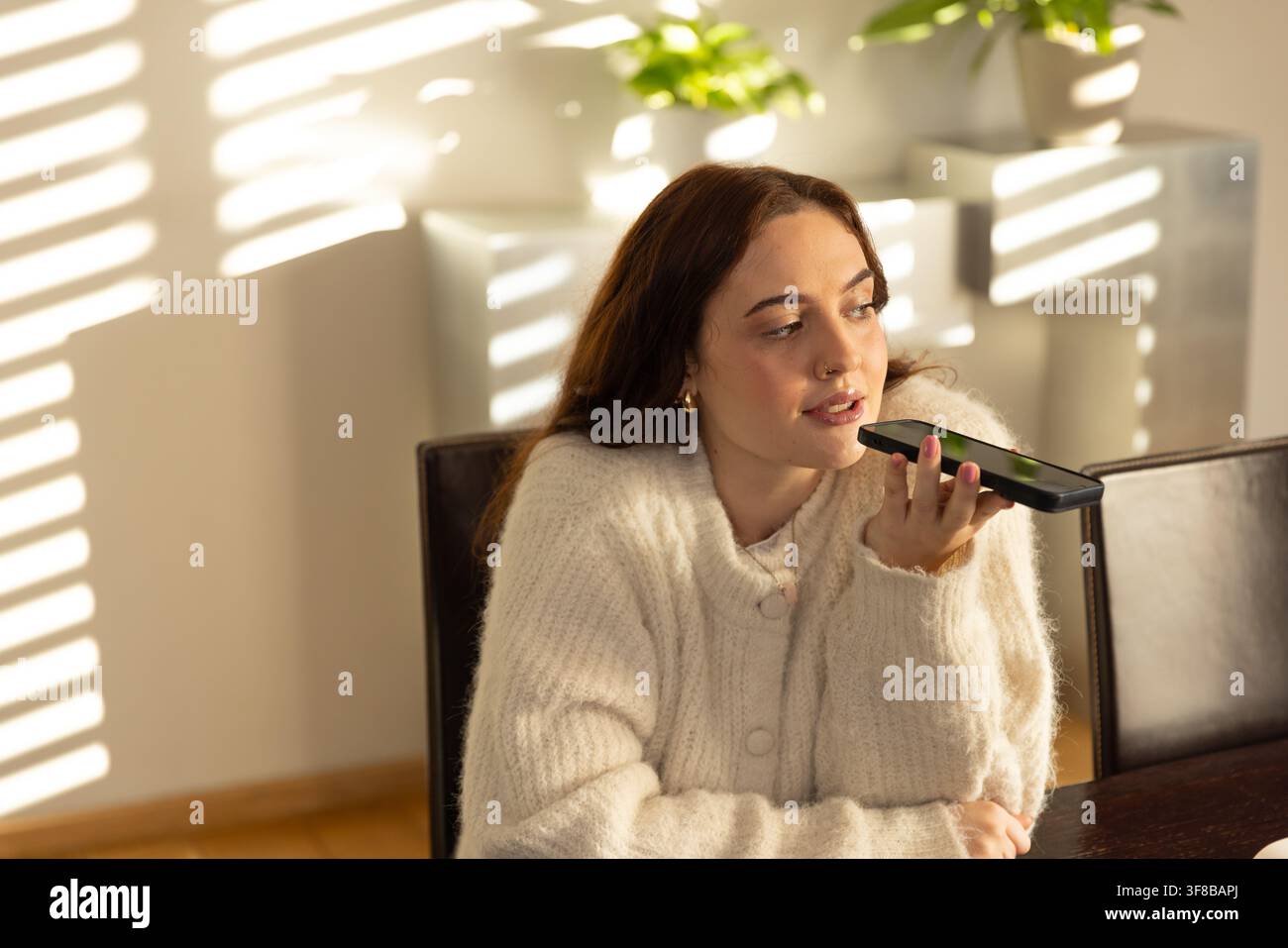 Frau an Holz-Esstisch spricht ins Telefon in cremefarbener Strickjacke, goldene Kreolen, Jalousien streifen die Wand. Weiblich, sonnenbeschienen, Pflanze, Leder, warm, gemütlich, Zuhause Stockfoto