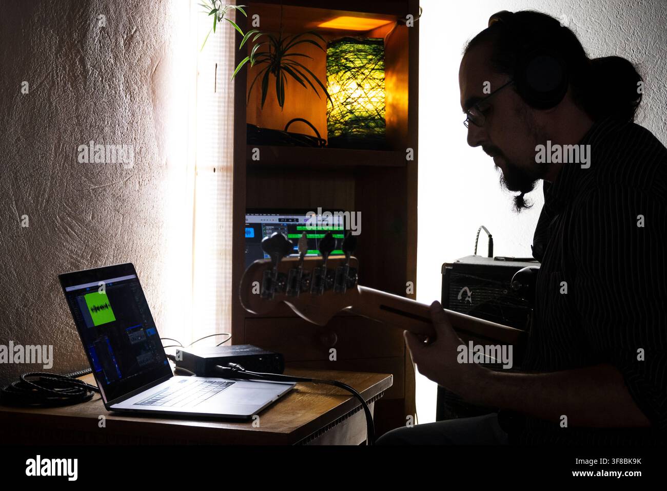 Kreativer Musiker, der in einem heimischen Musikarbeitsplatz vertieft ist, Bassgitarre spielt und einen Laptop sowie digitale Aufnahmesysteme verwendet. Stockfoto