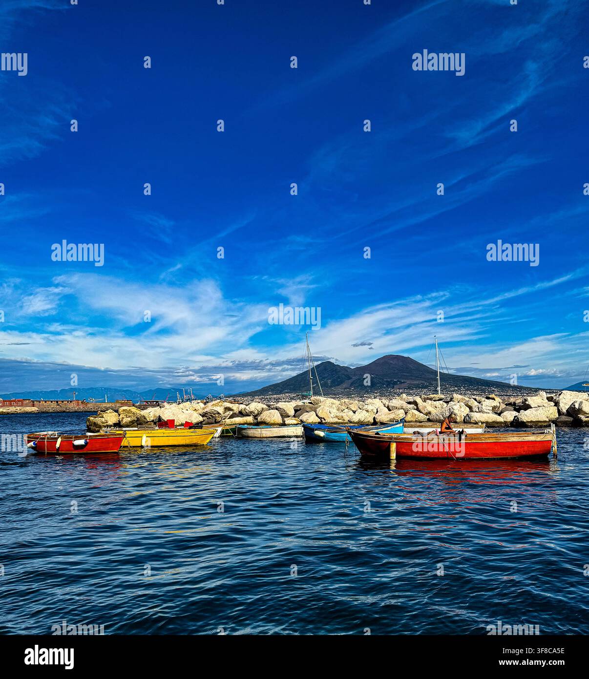 Boote in verschiedenen Farben sind auf dem Wasser in der Nähe eines felsigen Ufers zu sehen. Im Hintergrund ist unter einem blauen Himmel mit Wolken ein Berg sichtbar. - Smartphone-aufgenommenes Stockfoto