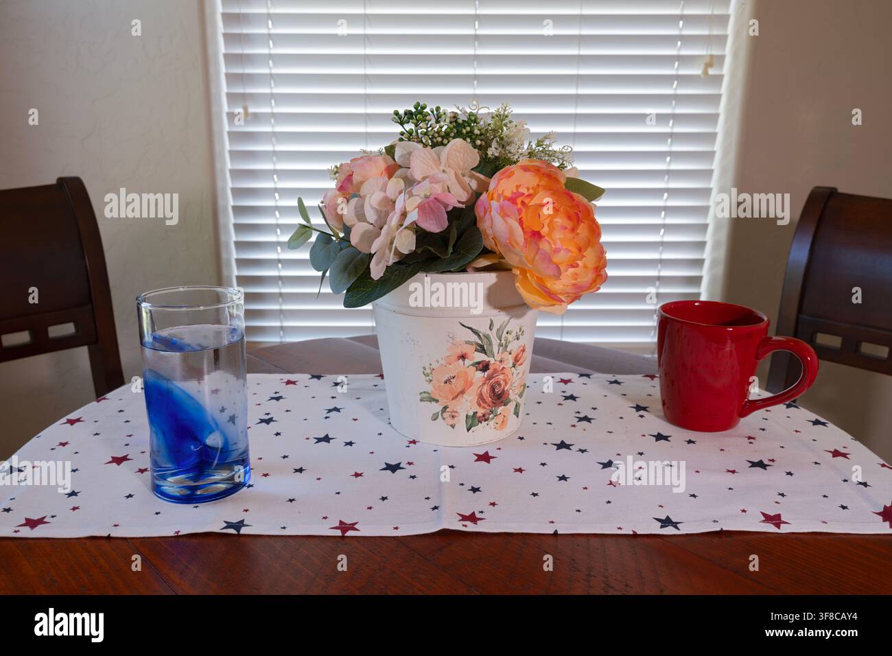 Tischgedeck bei einem Fenster mit einer Tasse, einem Glas und einem künstlichen Blumenmittelstück Stockfoto