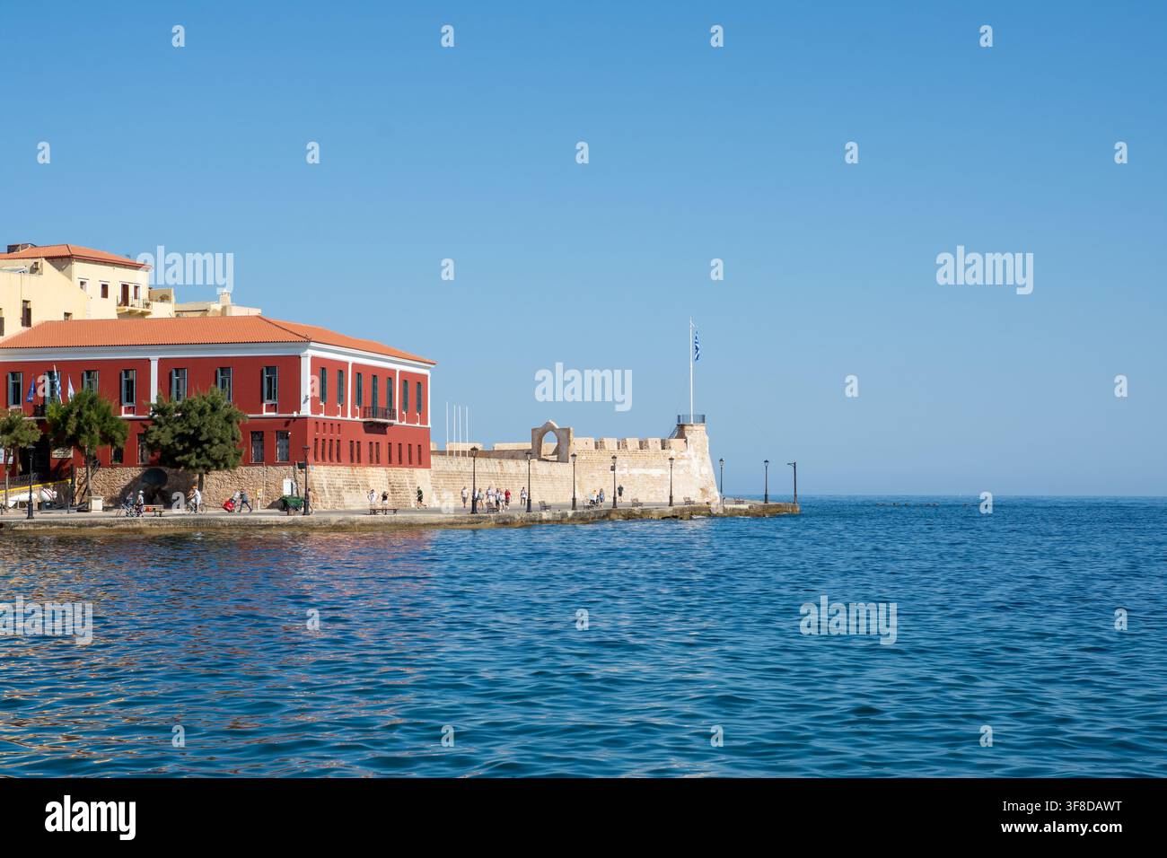 Blick auf das historische rote Gebäude und die Steinbefestigungen im Hafen der Altstadt von Chania mit Menschen, die am Ufer in Chania, Kreta, Griechenland spazieren gehen Stockfoto