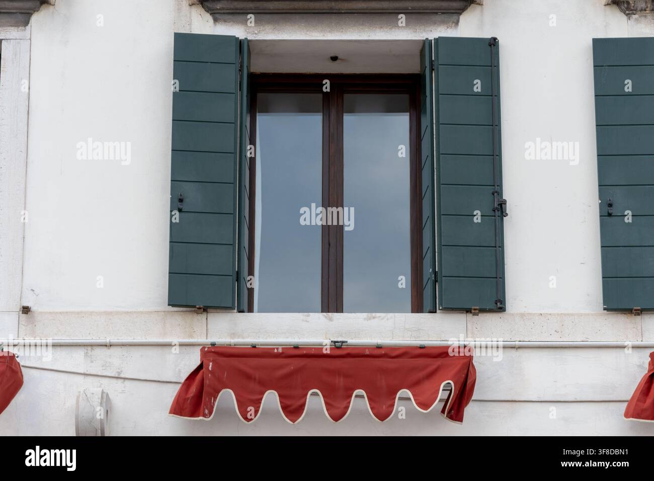 Ein hohes Glasfenster mit offenen grünen Fensterläden ist in einer weißen Fassade über einem roten Markisenvordach eingelassen. Stockfoto
