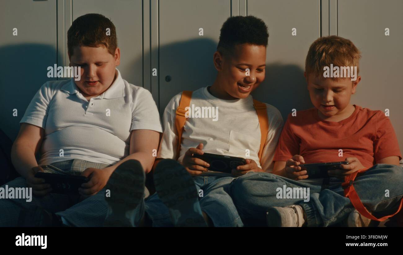 Grundschulklassenkameraden sitzen auf dem Flurboden mit Rucksäcken und digitalen Geräten, lächeln und verbringen Zeit zusammen vor dem Unterricht. Schulalltag, Freundschaft, Bildung und moderne Kindheit. Stockfoto