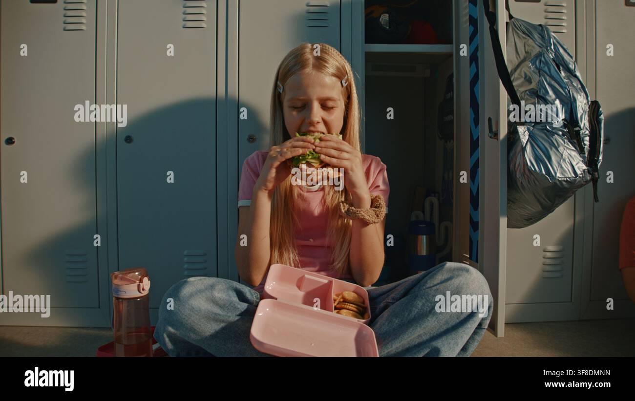 Blonde Grundschulmädchen genießt frisches Sandwich in rosa wiederverwendbarer Lunchbox, während sie neben Metallschließfächern in heller Schulflur sitzt. Ernährunggewohnheiten, Kindliche Unabhängigkeit, Moderne Bildung. Stockfoto