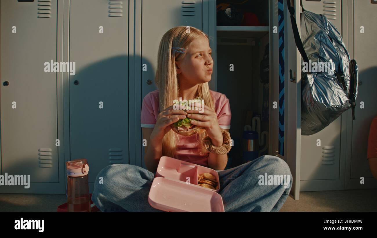 Blonde Grundschulmädchen genießt frisches Sandwich in rosa wiederverwendbarer Lunchbox, während sie neben Metallschließfächern in heller Schulflur sitzt. Ernährunggewohnheiten, Kindliche Unabhängigkeit, Moderne Bildung. Stockfoto