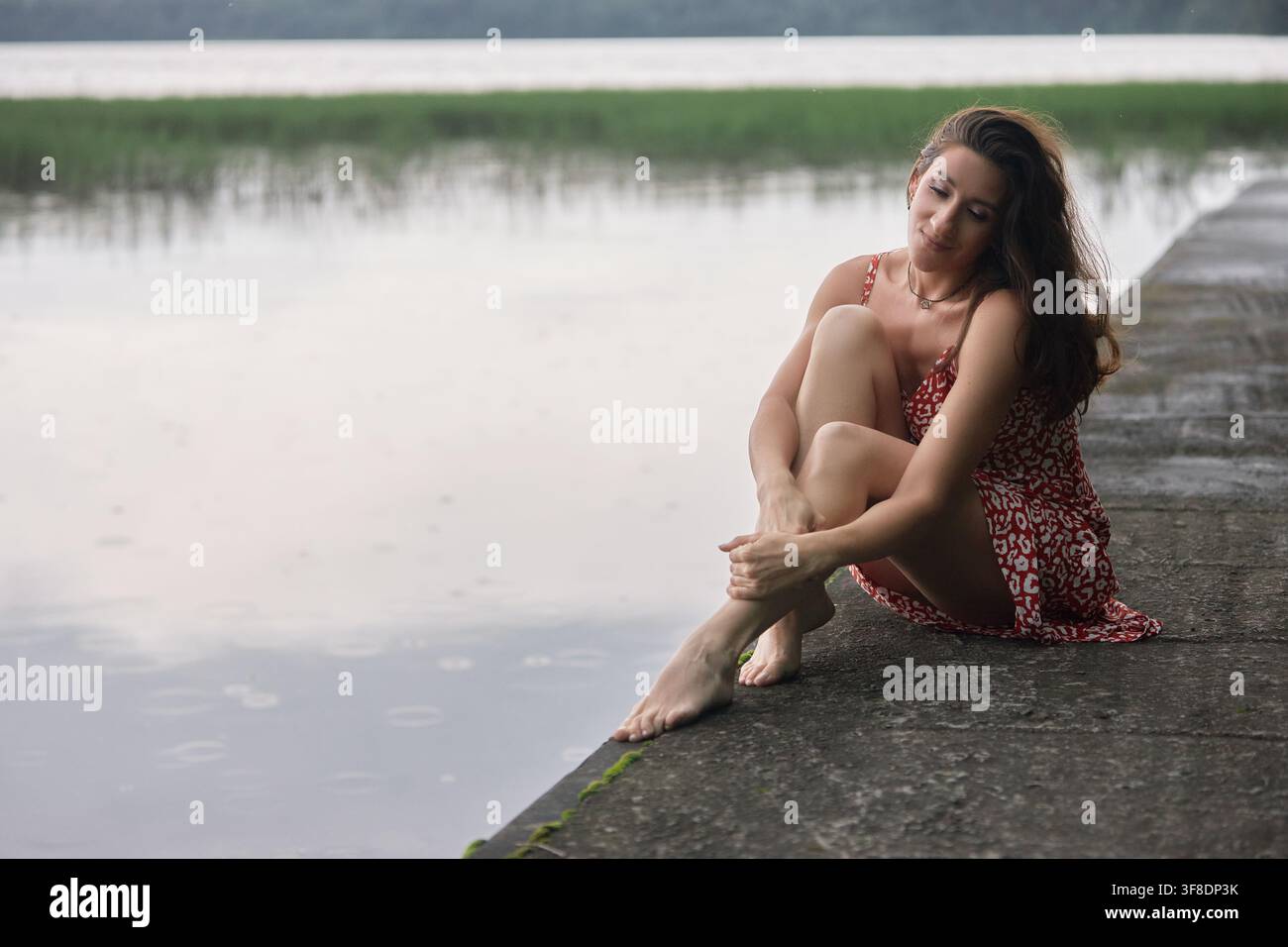 junge Frau sitzt auf einem steinernen Dock an einem Gewässer. Sie hat einen nachdenklichen Blick im Gesicht. Das Wasser ist ruhig, und der Himmel ist leicht bewölkt in der Nachmittagsstunde Stockfoto