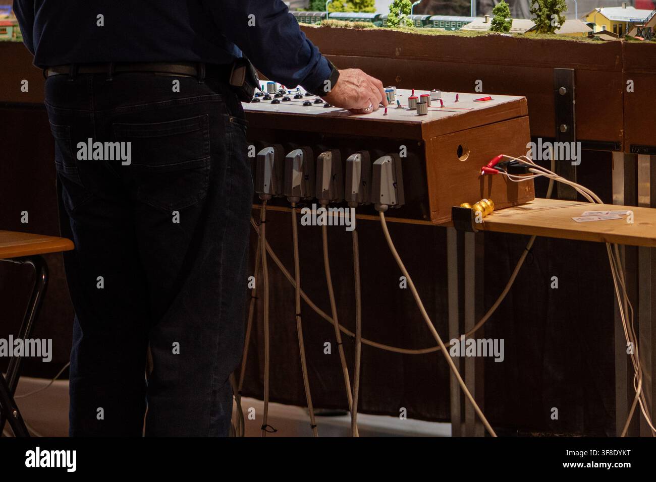 Ein erwachsener Mann steht neben einem hölzernen Bedienfeld mit Schaltern und Kabeln in einer indoor-Modellbahn-Anlage. Stockfoto