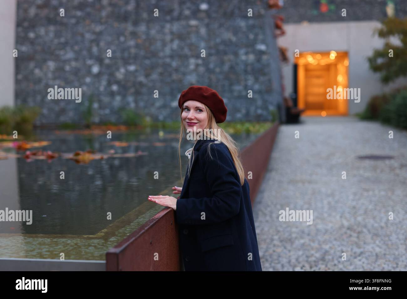 Modische junge Frau mit rotem Barett und schwarzem Mantel, die im Herbst in einer städtischen Umgebung am Wasser steht. Stockfoto