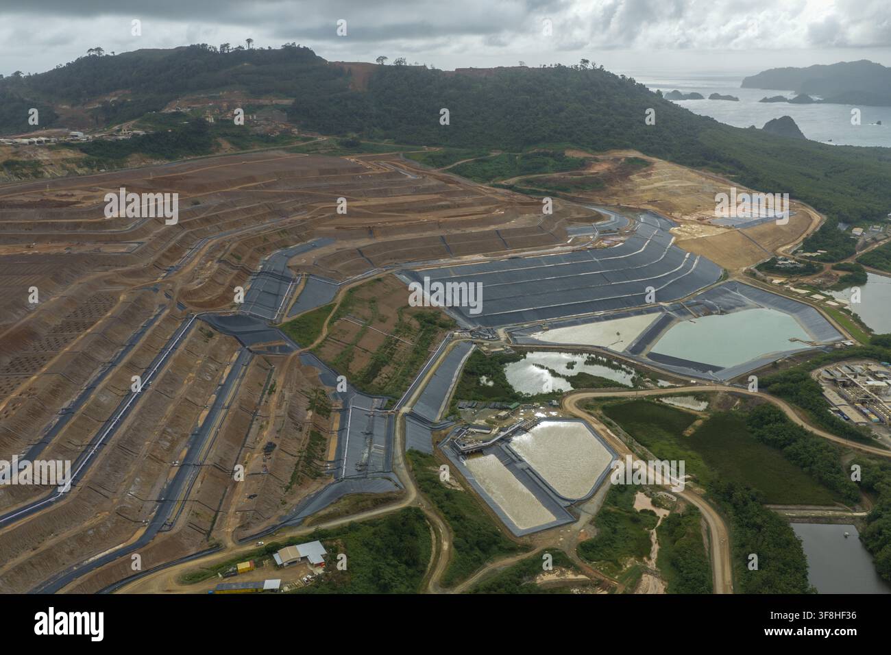 Luftaufnahme der großen Tagebau-Gold- und Kupfermine mit terrassierten Hängen, Sedimentteichen und den umliegenden üppigen Hügeln nahe der Küste in Java, Indonesien Stockfoto