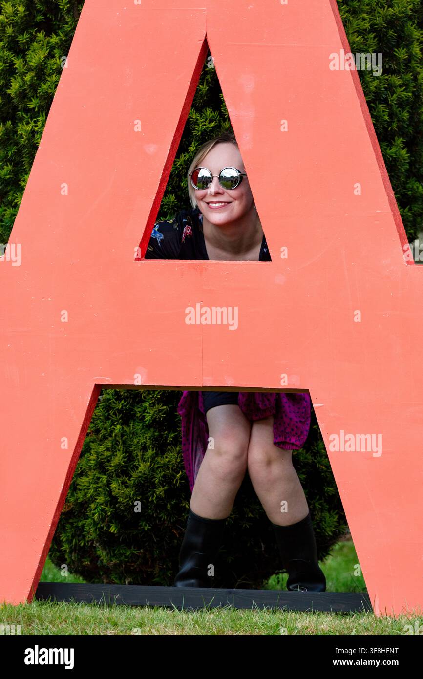 FRAU SCHAUT DURCH EINEN GIGANTISCHEN ROTEN BUCHSTABEN A, REPORTAGE, GREEN MAN FESTIVAL 2016, BRECON, WALES, UK, 2016: Eine lächelnde junge Frau mit blondem Haar und runden reflektierenden Sonnenbrillen schaut verspielt durch die dreieckige Öffnung eines großen, freistehenden roten Buchstabens „A“ im Freien. Foto: Rob Watkins. INFO: Das Green Man Festival begann 2003 in Wales. Jährlich findet es am dritten Wochenende im August in den Brecon Beacons statt und feiert alternative Musik, Kunst, Literatur, Wissenschaft und familienfreundliche Kultur in einer wunderschönen Berglandschaft. Stockfoto