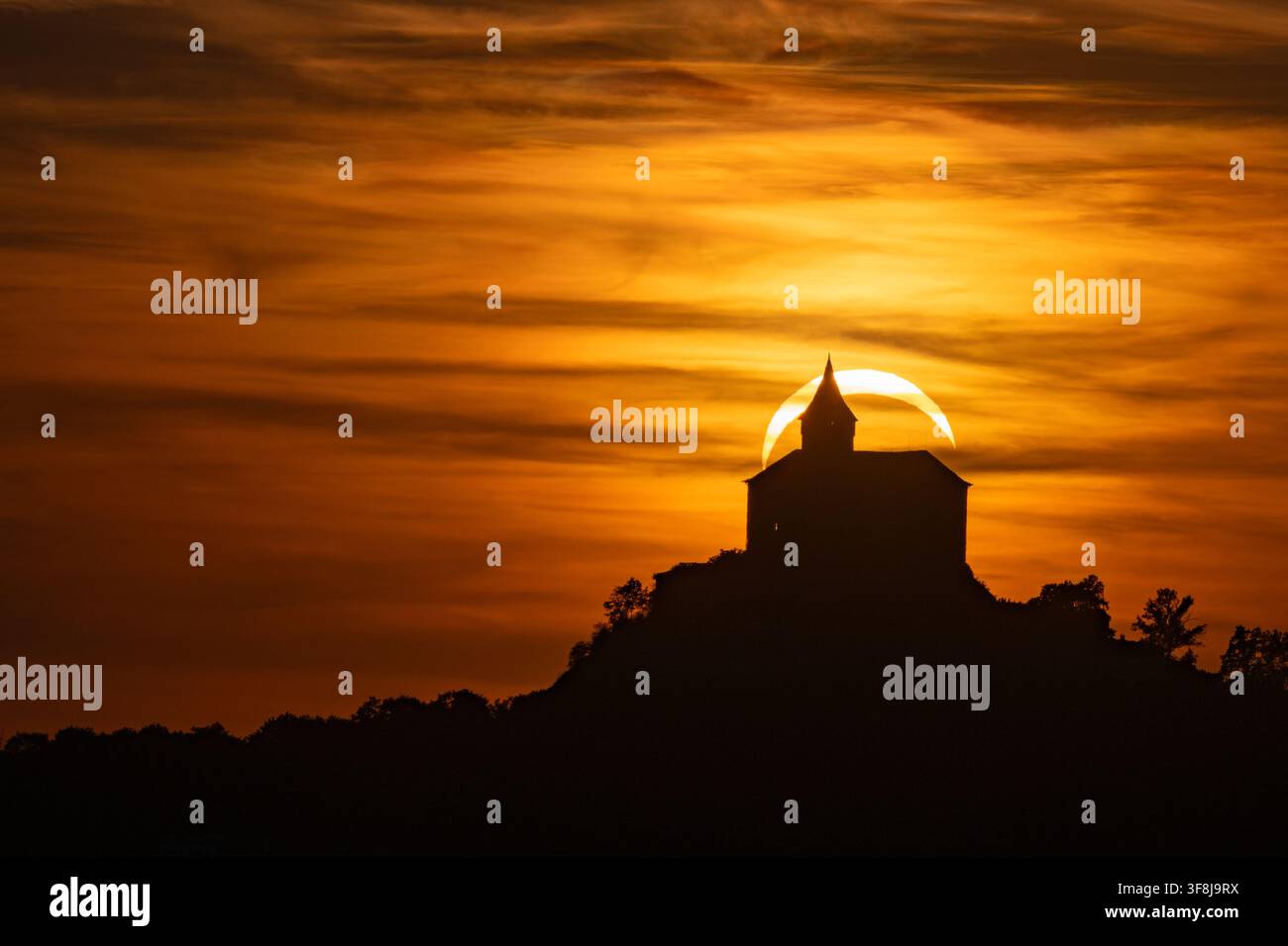 Teilweise Sonnenfinsternis im zunehmenden Mondstand, die sich direkt hinter der Silhouette des Kunetická hora Schlosses vor einem feurigen orangefarbenen Sonnenuntergangshimmel abzeichnet, Tschechische Republik. Stockfoto