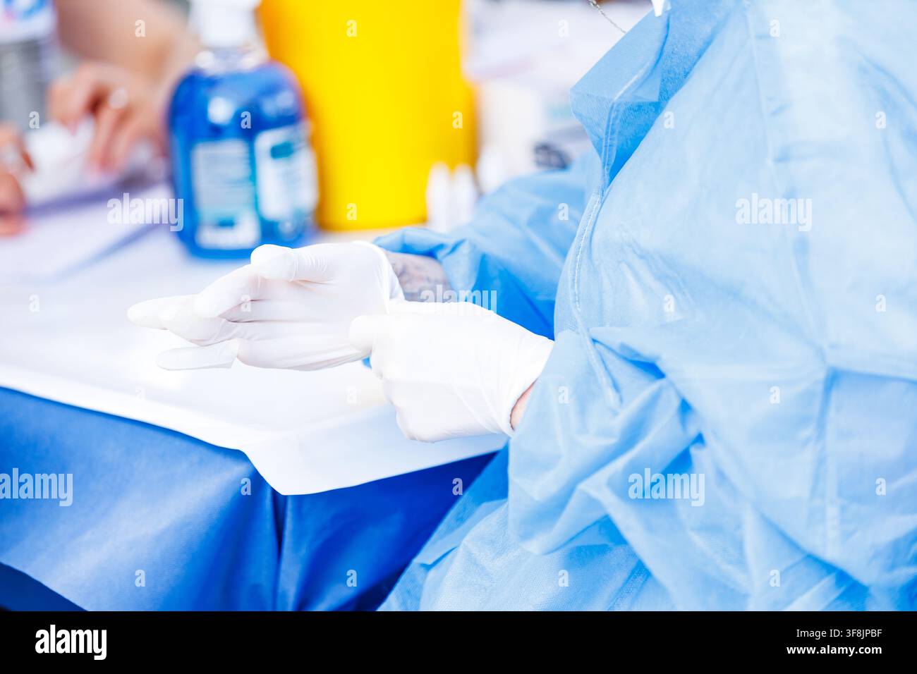 Nahaufnahme eines Gesundheitsfachkrafts in einem blauen Schutzkittel, der weiße sterile Latexhandschuhe anzieht, bevor er eine medizinische Prozedur oder einen klinischen Test durchführt Stockfoto