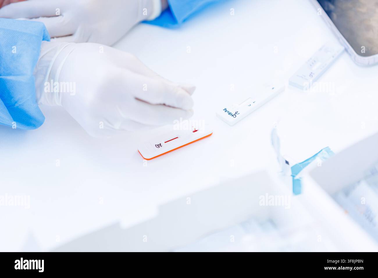 Nahaufnahme der Hände eines Gesundheitsfachkrafts in Schutzhandschuhen, der eine Blutprobe auf ein Schnelltestkassette auf einer weißen Oberfläche in einem medizinischen Umfeld aufträgt Stockfoto