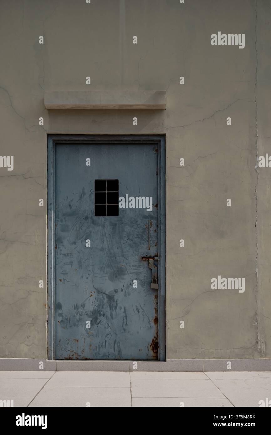 Rostige verschlossene Metalltür mit kleinem Fenster in rissiger Zementmauer. Stockfoto