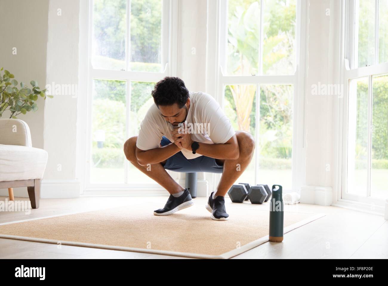 Mittelalter Afrikaner führt auf einer beigen Matte zu Hause in der Nähe von Hanteln und einer Wasserflasche einen tiefen Kniebeugen durch Stockfoto