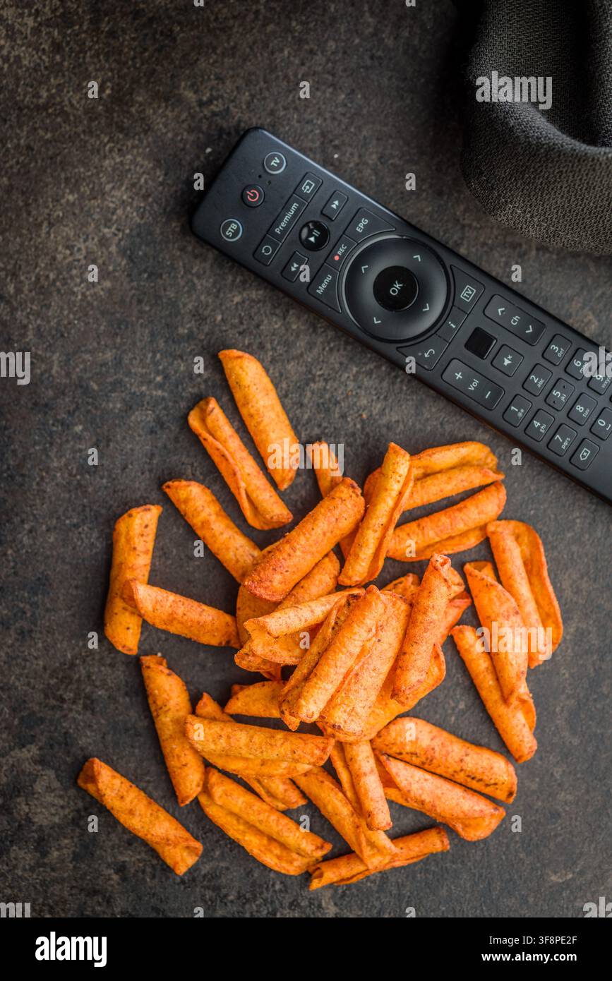 Scharfe knusprige Mais-Snack-Stäbchen, die neben einer TV-Fernbedienung auf einer dunklen Oberfläche verstreut sind, was auf lässiges Filmabend-Snacking, Komfortessen-Genuss und Entspannung hindeutet Stockfoto