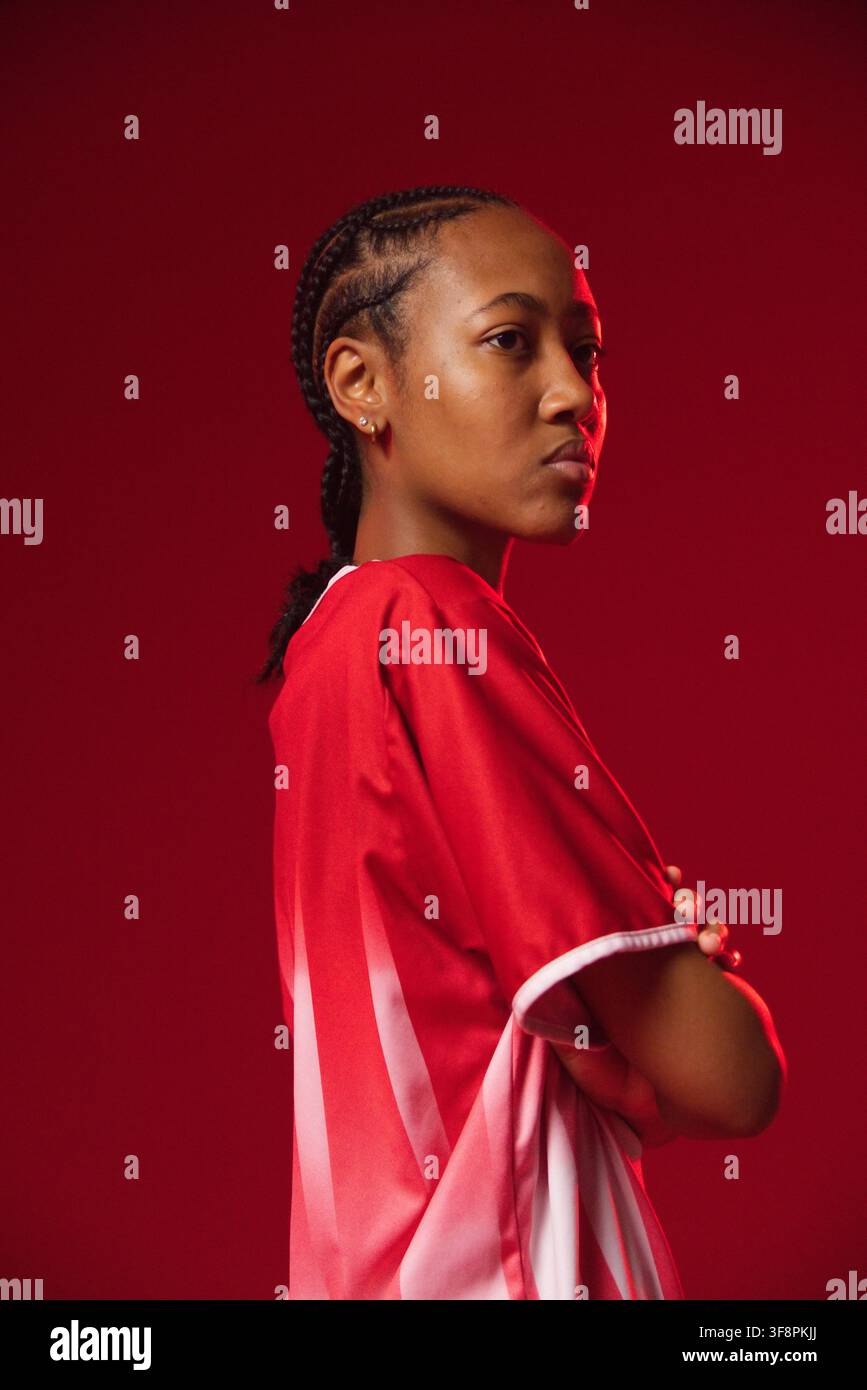 Afroamerikanische Frau im Profil im Studio posiert, trägt rotes-weißes Sporttrikot, Zöpfe, Kreolen Stockfoto