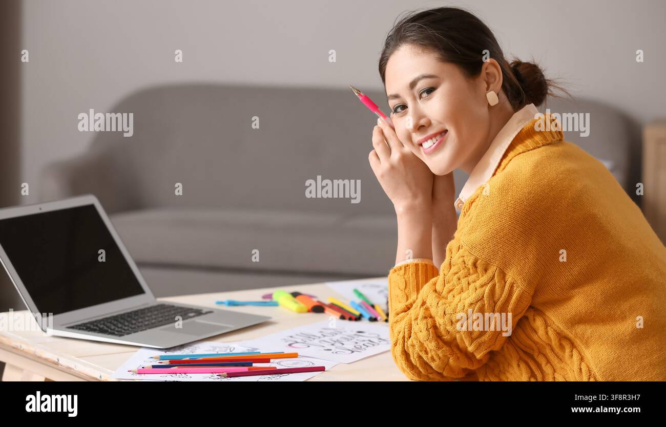 Lächelnde junge asiatische Frau, die einen rosa Stift hält, während sie an einem Schreibtisch mit einem Laptop und Kunstutensilien an kreativen Projekten arbeitet Stockfoto