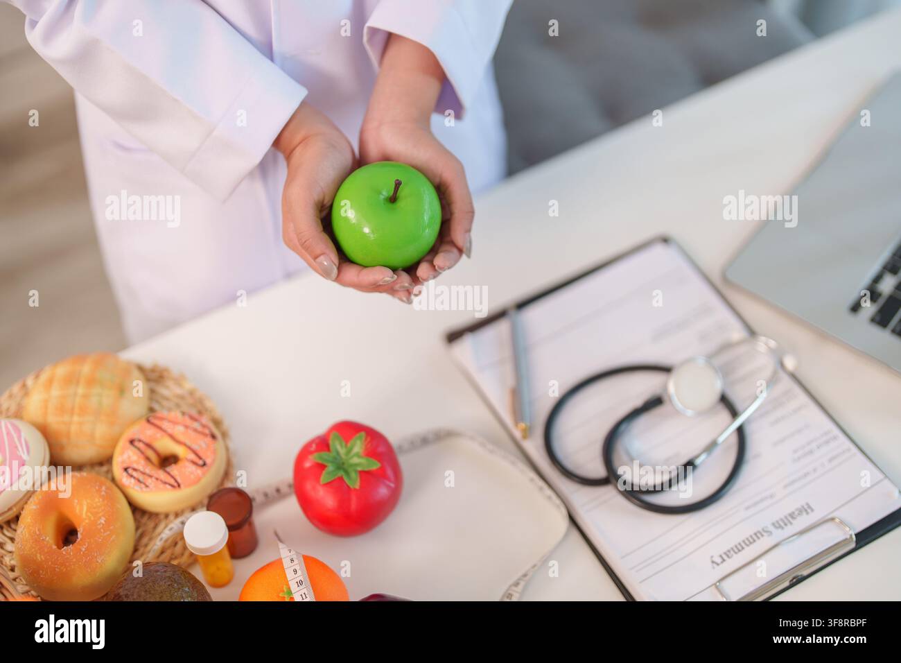 Hochwinkelsicht einer Ernährungsberaterin, die einen grünen Apfel über einem Stethoskop und einer Gesundheitszusammenfassung hält Stockfoto