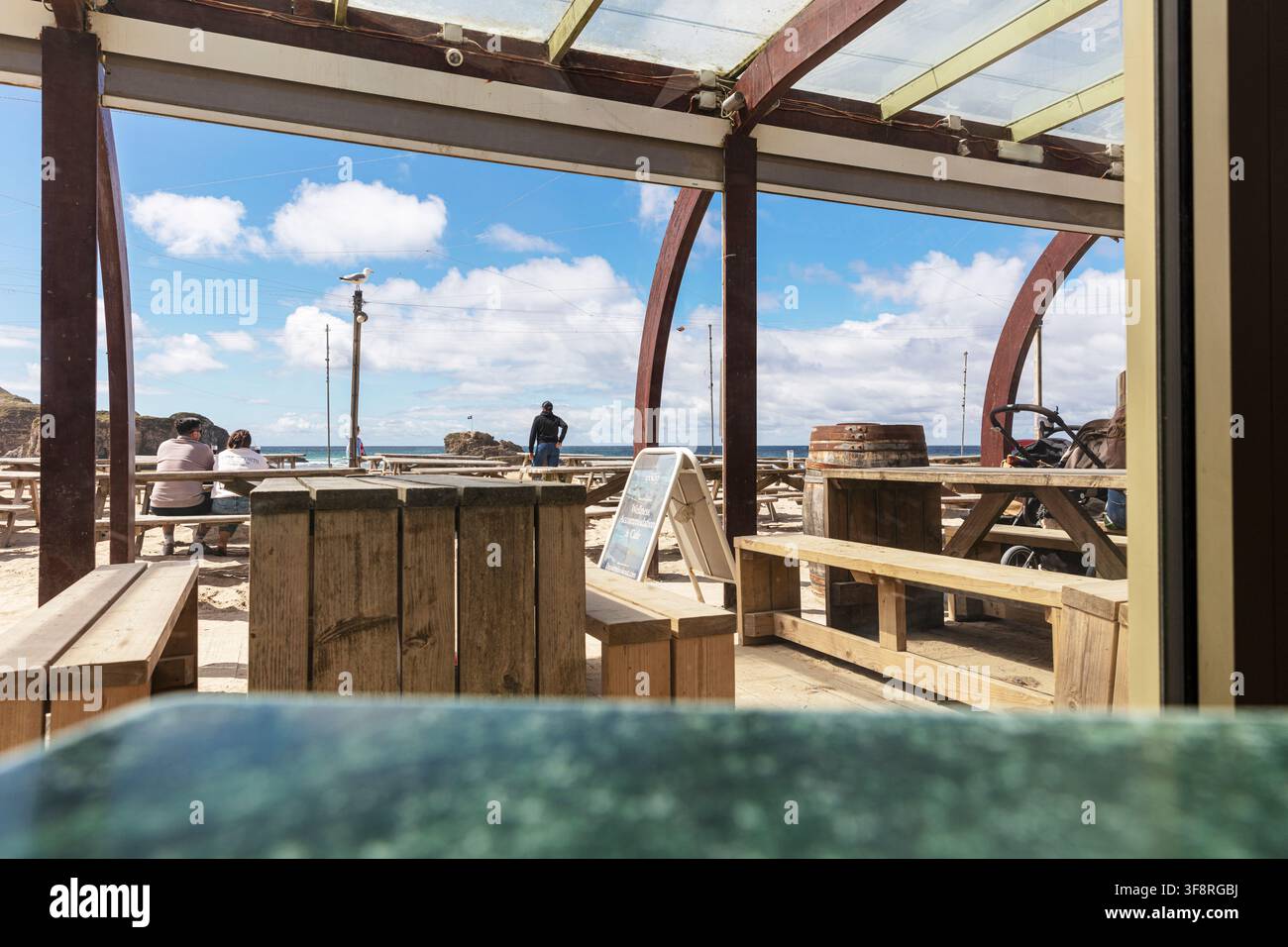 Der Wasserloch, Perranporth, Cornwall, UK, England. Außensitzgelegenheiten mit Holzbänken und einem gläsernen Pavillon, der den Ozean dahinter einrahmt. Stockfoto