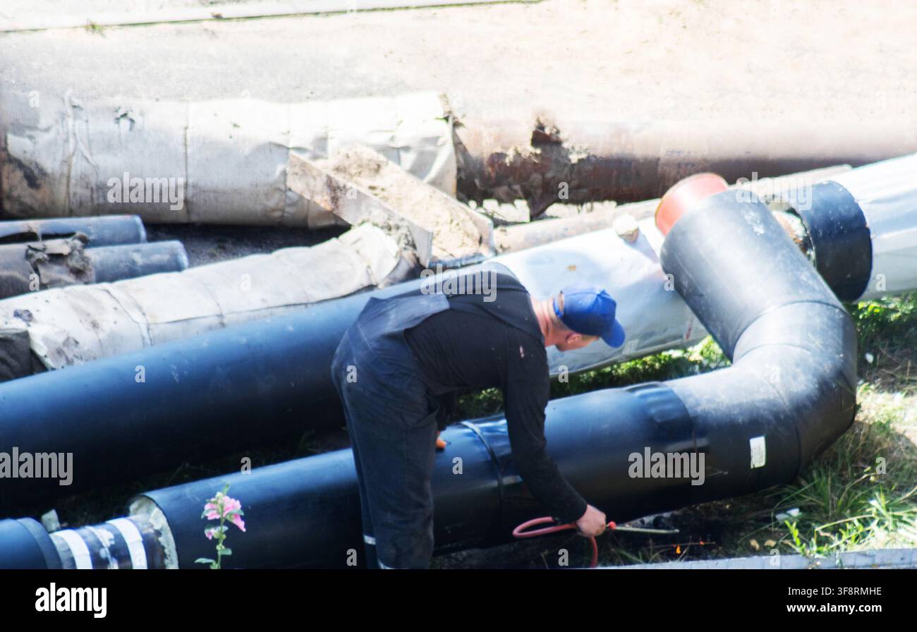 Erwachsener Arbeiter in blauer Kappe und dunkler Uniform, der aktiv am Verbindungsprozess eines neuen Heizrohrsystems beteiligt ist. Die Arbeit umfasst Schwarze Rohre mit großem Durchmesser Stockfoto