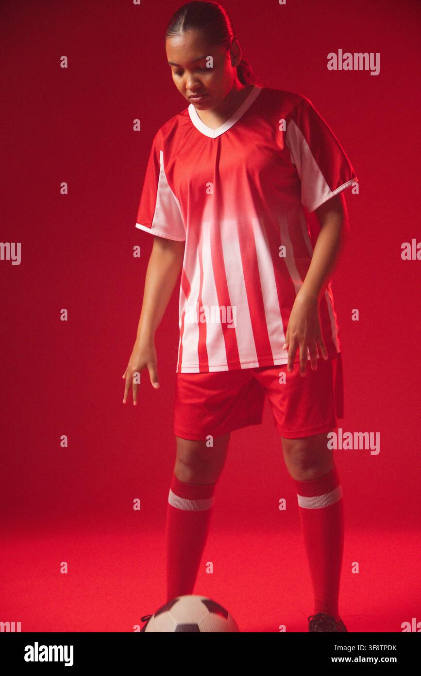 Afroamerikanerin steht auf rotem Studioboden, trägt ein rot gestreiftes Trikot und schaut auf den Ball Stockfoto