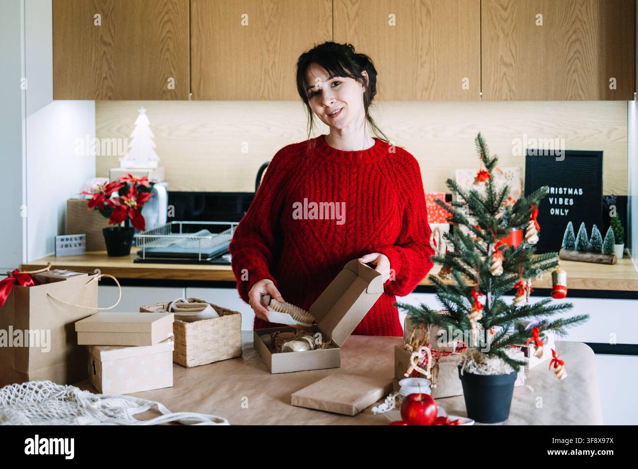 Frau hält hölzernen Pinsel über offene Kraftbox in der Küche in der Nähe des Schildes „CHRISTMAS VIBES ONLY“. Bereitet Weihnachtsbestellung zur Versendung vor, montiert Produkt für Stockfoto