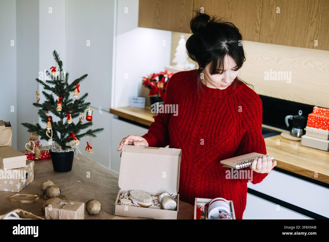 Frau hält Notizblock und überprüft eine offene Kiste mit Pinsel und Glas in der Küche. Kleinunternehmen überprüft die Bestellliste, bestätigt den Inhalt der Weihnachtsbestellung Stockfoto