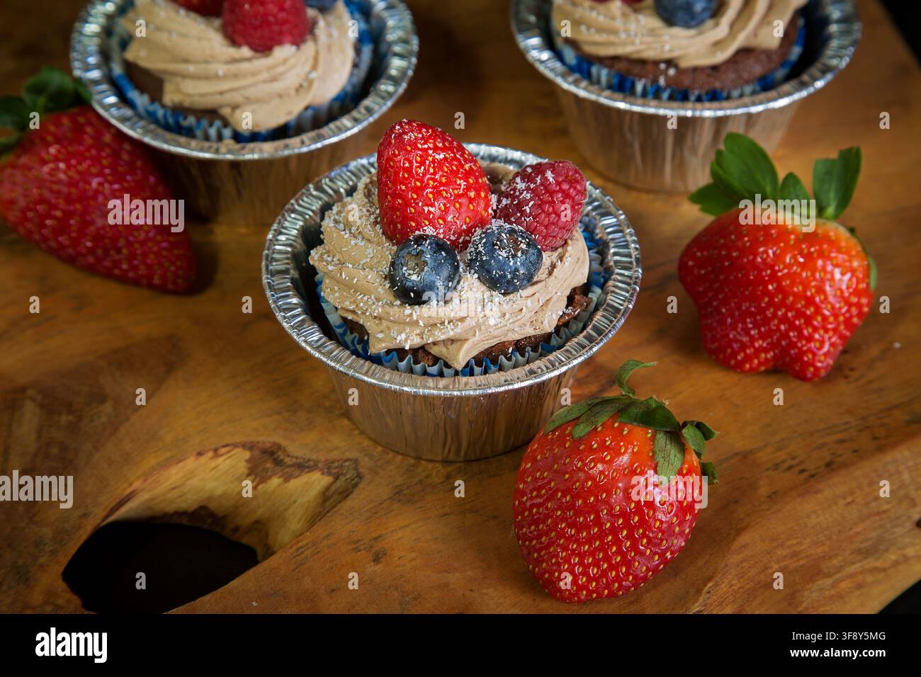Schokoladen-Cupcakes, die mit Erdbeeren, Blaubeeren und Himbeeren auf einem Holzbrett auf schwarzem Hintergrund dekoriert sind. Hochwertiges Foto Stockfoto