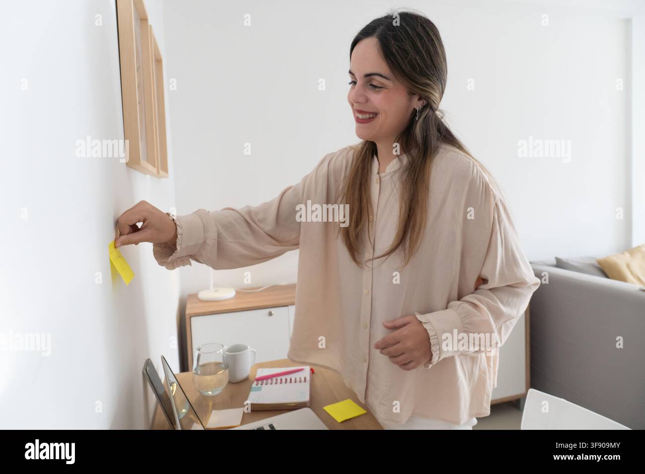 Lächelnder Content-Ersteller platziert einen gelben Klebezettel an der Wand neben einem gerahmten Druck, Schreibtisch mit Laptop und Notizbuch sichtbar in einer hellen Wohnung Stockfoto