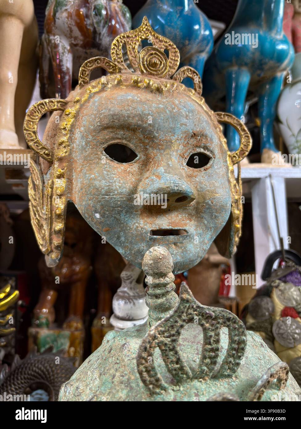 Mercado de las Pulgas in Buenos Aires, Argentinien. Asiatische Ritual-Bronzemaske mit grüner Patina, vergoldeten Kronendetails und Keramikskulpturen im Hintergrund Stockfoto
